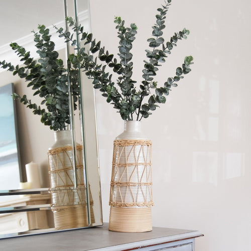 Two tone ribbed vase with rattan sleeve and eucalyptus on table.